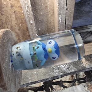 May include: A clear glass tumbler with a blue and white cartoon character inside. The character has a blue head with a yellow accent and white eyes. The glass is resting on a wooden surface.