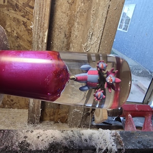 May include: A clear glass object with a vibrant magenta interior and a decorative, colorful, cartoon-like design. The object is mounted on a wooden lathe, showcasing the craftsmanship and design.