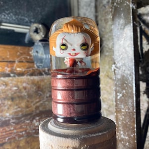 May include: A clear glass bottle with a red and white figure inside. The figure is a clown with a red nose and yellow eyes. The bottle is sitting on a wooden surface.