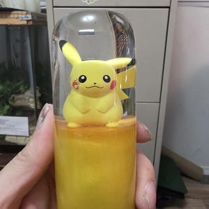 May include: A clear, cylindrical container holding a yellow Pikachu figure. The container is filled with a shimmering yellow liquid. The Pikachu figure has black-tipped ears, red cheeks, and a smiling expression. The container is held in a hand.