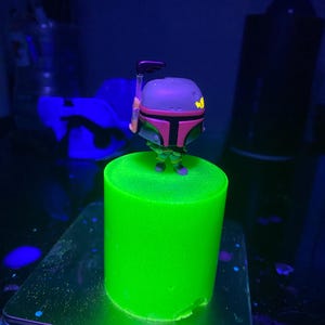 May include: A green glow-in-the-dark Boba Fett figurine stands on a green glow-in-the-dark cylinder. The figurine is holding a weapon.
