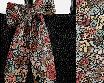 Pode incluir: Uma mala de mão preta com acabamento texturizado e alça preta, adornada com uma fita de tecido comprida atada com um padrão floral denso em tons de vermelho, amarelo, castanho e verde sálvia sobre fundo preto, visível em detalhe à direita.