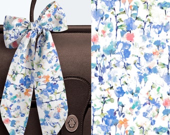 May include: A white silk ribbon tied in a bow featuring a watercolor floral print in shades of blue, green, and coral. The ribbon is draped over a dark brown leather bag with a metallic clasp, and a detailed view of the floral pattern is shown.