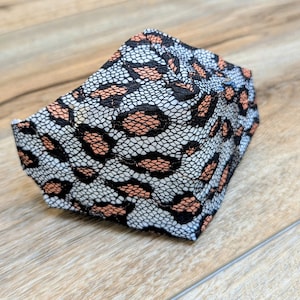 May include: A folded face mask featuring a leopard print design. The mask has a white background with black and peach-colored spots. The mask is made of a lace-like material.