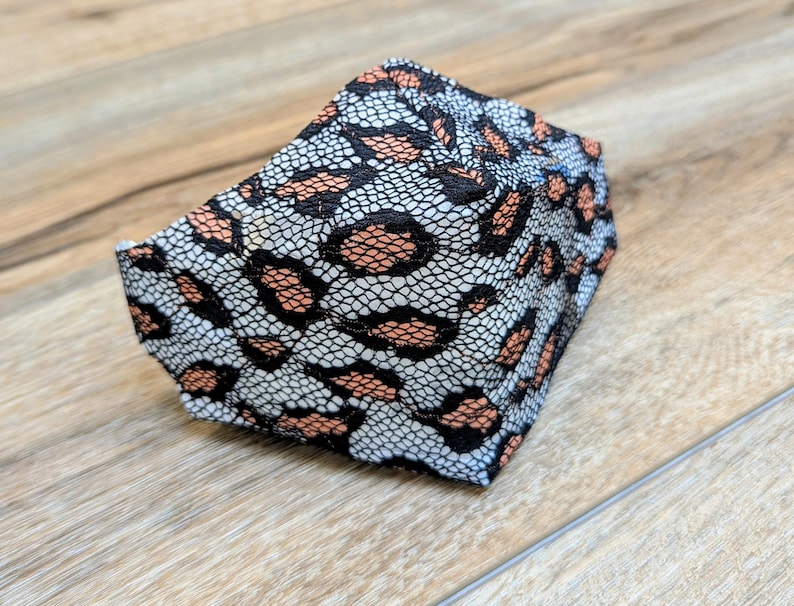 May include: A folded face mask featuring a leopard print design. The mask has a white background with black and peach-colored spots. The mask is made of a lace-like material.