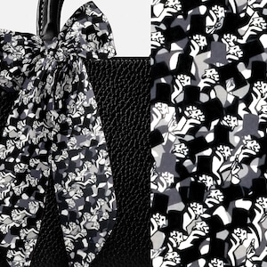 May include: A black textured handbag adorned with a long, tied ribbon scarf featuring a repeating graphic pattern of black, white, and gray top hats and abstract shapes. The scarf is tied in a bow around the handle of the structured leather bag.