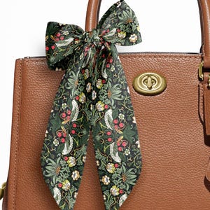 May include: A brown pebbled leather handbag featuring a brass turn-lock closure and a hanging leather tag. A long, thin scarf with a dark green background and a repeating pattern of white birds, red berries, and yellow flowers is tied to the handle.