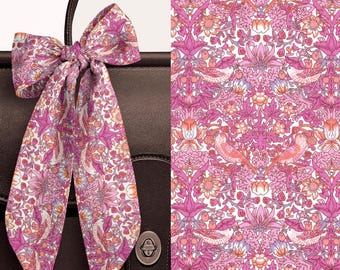 May include: A long, thin silk scarf tied in a bow, featuring a repeating pattern of pink and white birds, flowers, and foliage. The scarf is displayed against a dark brown leather bag, with a separate panel showing the detailed floral bird print.