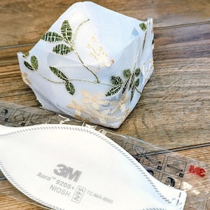 May include: A white face mask with gold and green floral embroidery sits atop a wooden surface. Below it is a 3M Aura 9205+ N95 respirator mask in a clear plastic package.