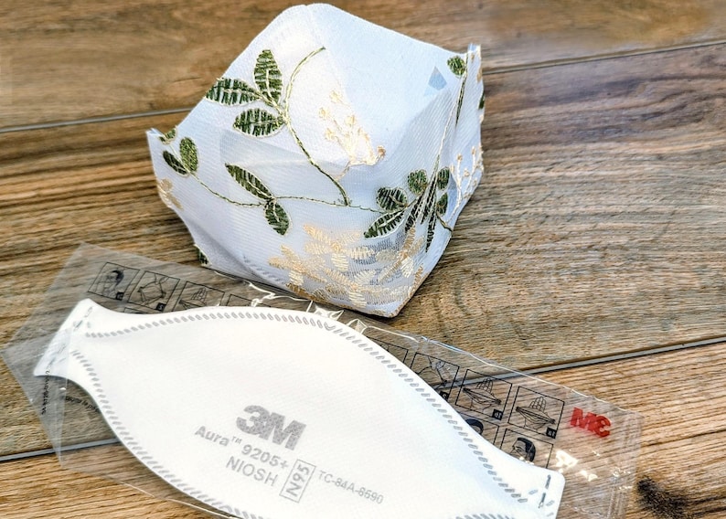 May include: A white face mask with gold and green floral embroidery sits atop a wooden surface. Below it is a 3M Aura 9205+ N95 respirator mask in a clear plastic package.