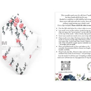 May include: White reusable face mask cover with a floral pattern of pink flowers and green stems. The mask cover has the text "M" and other text. The mask cover is designed to add style and color to your Aura mask.