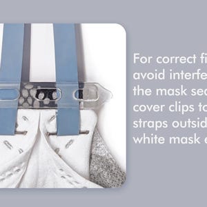 May include: A white mask with blue straps and a clear plastic clip. The text on the right side of the image reads: "For correct fit and to avoid interfering with the mask seal, mask cover clips to the straps outside the white mask edge."