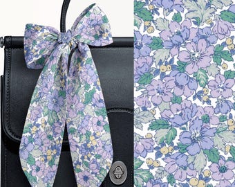 May include: A long, thin fabric ribbon tied in a bow, featuring a floral pattern with light purple flowers, green leaves, and small yellow berries on a white background, draped over the top handle of a black leather handbag.