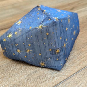 May include: A blue fabric item with a celestial pattern of gold stars and dots. The item is a geometric shape, possibly a box or a pouch, and is resting on a wooden surface.