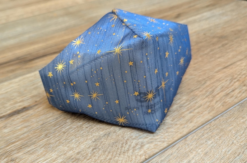 May include: A blue fabric item with a celestial pattern of gold stars and dots. The item is a geometric shape, possibly a box or a pouch, and is resting on a wooden surface.