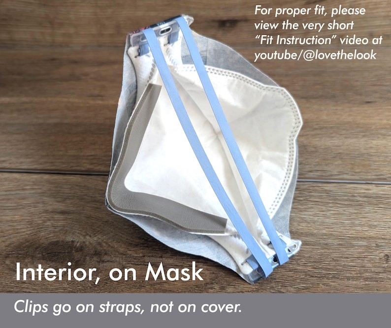 May include: A white and gray mask with light blue straps. The mask has the text "Interior, on Mask" and "Clips go on straps, not on cover." The image also includes the text "For proper fit, please view the very short "Fit Instruction" video at youtube/@lovethelook."