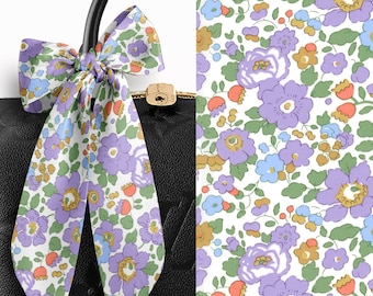 May include: A long, thin silk ribbon tied in a bow around the handle of a black textured handbag. The ribbon features a white background with a repeating floral pattern in shades of purple, light blue, green, and orange. A gold padlock is visible.