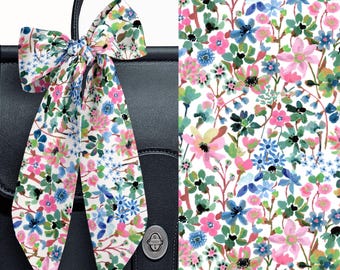 May include: A long, thin fabric scarf tied in a bow, featuring a white background with a watercolor floral print in shades of pink, blue, and green. The scarf is draped over a dark navy blue leather bag with a silver-toned metal turn-lock closure.