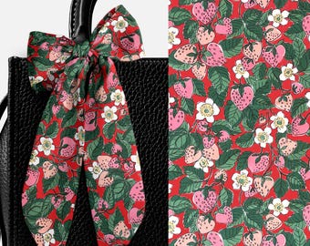 Pode incluir: Uma fita de tecido longa e estreita atada em laço, com um fundo vermelho vibrante e um padrão repetitivo de morangos cor-de-rosa, folhas verdes e flores brancas. A fita está presa à alça de uma mala de mão preta texturizada.