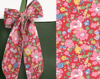 May include: A dark green textured leather tote bag adorned with a long, tied fabric ribbon featuring a vibrant red background with a repeating pattern of pink flowers, yellow blossoms, light blue buds, green leaves, and small gold-toned stars.