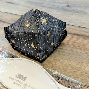 May include: A black face mask with a gold star pattern. The mask is folded and sits on a wooden surface next to a white filter with the text "3M Aura 9205+".