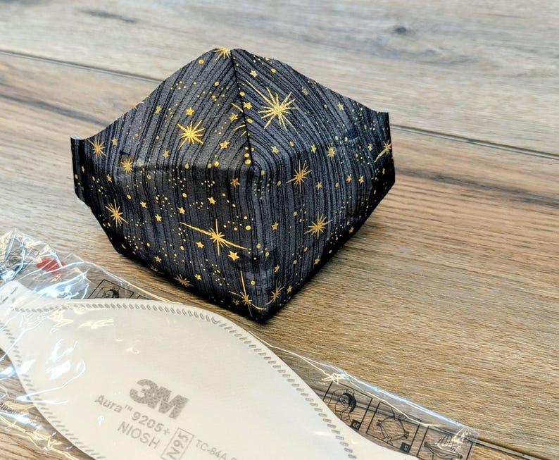 May include: A black face mask with a gold star pattern. The mask is folded and sits on a wooden surface next to a white filter with the text "3M Aura 9205+".