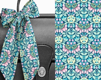 May include: A long, narrow silk scarf tied in a bow, featuring a repeating floral pattern with pink, light green, and yellow blossoms against a dark blue background. The scarf is draped over a black leather bag with a silver metal turn-lock closure.