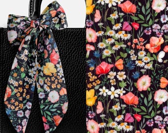 May include: A black textured handbag featuring a long, silky scarf tied in a bow around the handle. The scarf displays a vibrant floral pattern on a black background, showcasing various flowers in shades of red, yellow, white, and blue.