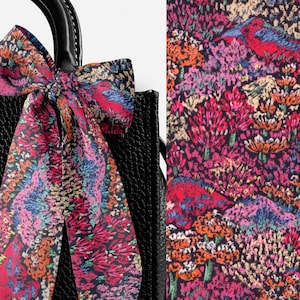 May include: Black tote bag with a floral scarf tied to the handle. The scarf features a vibrant pattern of red, pink, blue, and orange flowers and foliage. The bag has a textured surface.