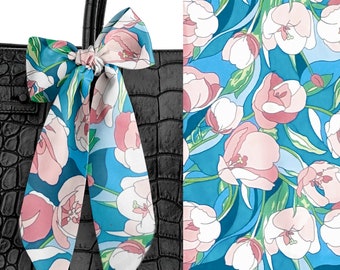 May include: A long, narrow silk scarf tied in a bow on a black crocodile-textured handbag. The scarf features a repeating pattern of light pink tulips with green leaves against a vibrant blue background, shown both tied and as a flat fabric swatch.