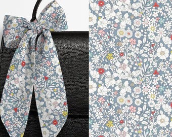 May include: A long, narrow fabric ribbon tied in a bow, featuring a dense floral pattern with white, pink, yellow, and red flowers on a dusty blue background, attached to the handle of a black textured handbag with a flap closure.