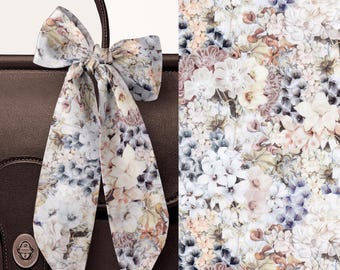 May include: A long, thin silk scarf tied in a bow, featuring a detailed floral print with shades of white, soft blue, and muted brown. The scarf is draped over a dark brown leather bag with a metallic turn-lock closure visible at the bottom.