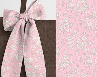 May include: A dark brown textured tote bag featuring a long, light pink ribbon tied into a bow at the handle. The ribbon displays a repeating pattern of small white flowers and leaves, which is also shown in a larger flat view on the right side.