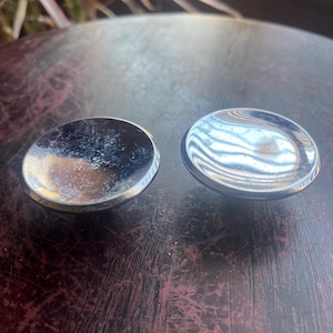 May include: Two silver-colored metal knobs with a rounded, slightly domed top. The knobs are sitting on a dark brown wooden surface.