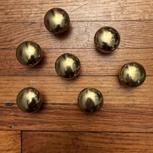 May include: A collection of six spherical brass knobs with a polished, reflective gold-tone finish. Each knob features a smooth, rounded surface and a consistent circular shape, arranged in a scattered pattern across the frame.