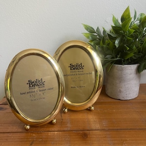 May include: Two oval, gold-colored brass photo frames with a polished and lacquered finish. Each frame has a stand and the text "Solid Brass PHOTO FRAME" and measurements of 3.5" x 5" (9cm x 13cm) and 3.5" x 5" (9cm x 13cm).