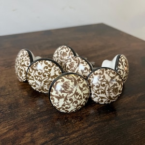 May include: A collection of seven ceramic cabinet knobs. Each knob features a cream-colored background with a brown floral pattern. The knobs have a black rim and a silver screw. The knobs are arranged on a dark brown wooden surface.