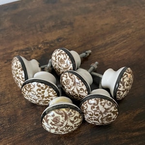 May include: A collection of seven ceramic cabinet knobs. Each knob features a cream-colored base with a brown floral pattern, a black border, and a silver screw. The knobs are arranged on a dark brown wooden surface.