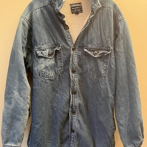 Puede incluir: Chaqueta vaquera azul con forro crema y difuso. La chaqueta tiene dos bolsillos en el pecho con cierres de botón y una parte delantera abotonada. El cuello es de estilo clásico y la chaqueta parece estar bien usada.