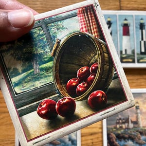 May include: A square ceramic coaster featuring a printed illustration of a wooden bucket tipped over on a windowsill, spilling bright red apples. The scene includes a view of a green tree through the window and a red and white chequered curtain.