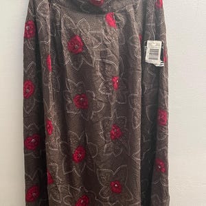 May include: A brown midi skirt featuring a repeating pattern of red flowers with white dotted outlines. The skirt has a high waistband and is displayed on a black hanger. A small white retail tag is attached to the upper right side of the garment.