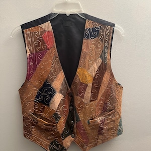 May include: A sleeveless vest featuring a patchwork design of various brown, tan, and dark-colored fabric segments. The front panels are decorated with intricate gold-toned swirling embroidery patterns and include two front pockets and a button closure.