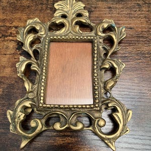 May include: Ornate brass picture frame with a rectangular opening and intricate floral and scrollwork details. The frame has a warm, aged patina and a small, decorative top element. The frame is designed to hold a small photograph or artwork.