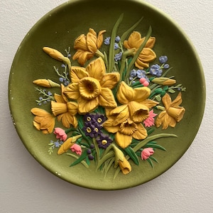 May include: A round olive green decorative plate featuring a raised, three-dimensional relief of yellow daffodils, small purple flowers, and tiny pink and blue blossoms with green leaves and stems arranged in a central floral bouquet pattern.