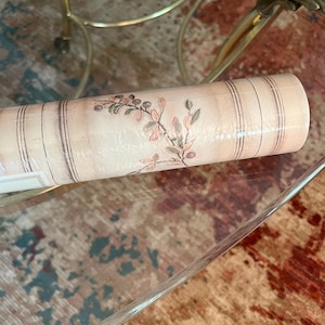 May include: A roll of wallpaper featuring a floral design with pink and green leaves and stems. The wallpaper has a light pink background with vertical brown stripes. The roll is wrapped in clear plastic.