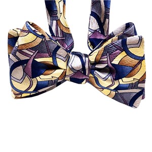 May include: A patterned bow tie featuring a geometric design in shades of blue, yellow, purple, and brown. The bow tie is a classic accessory, perfect for formal events or adding a touch of style to any outfit.