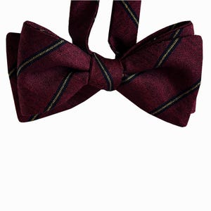May include: A deep burgundy bow tie featuring a textured fabric finish and thin, diagonal stripes in dark navy blue and gold. The bow tie is pre-tied with a central knot and includes an adjustable fabric neck strap extending from the top center.