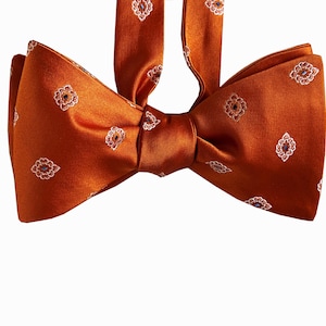May include: A burnt orange bow tie featuring a repeating pattern of small, white-outlined floral motifs with blue and red centers. The fabric has a smooth, satin-like sheen, and the bow tie includes an adjustable neck strap in the same material.