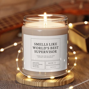 Puede incluir: Una vela marrón con una etiqueta blanca que dice "Smells Like World's Best Supervisor" y "Thank you for all of your support and guidance!" La vela está encendida y se encuentra sobre un posavasos de madera.