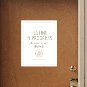 Può includere: Un cartello bianco con testo nero che recita "Testing in Progress, please do not disturb" con un'icona a forma di mano. Il cartello è su una porta di legno.